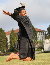 A photo Patricia in black graduation attire, jumping midair while smiling and looking at the camera outside in a grassy field.