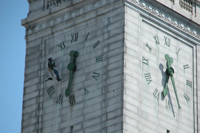 A man is suspended on wires from the top of the Campanile and is actively removing one of the clock hands. 