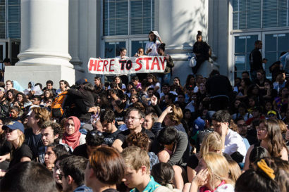 A photo of a crowd in protest in front of Sproul Hall with the person at the top of the steps holding a sign that reads "here to stay".