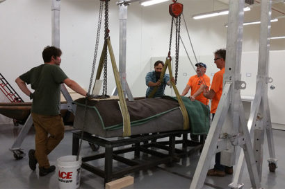 A photo of the four men standing around a casket that is attached to two ropes that loop around the upper and lower halves in a room with all white walls, ceiling, and lights.