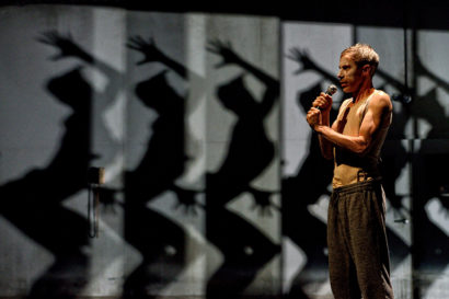 A photo of Kidd Pivot standing on stage with a microphone in front of a background that has six silhouettes lined up in an upward and bent pose with an arm above their heads.