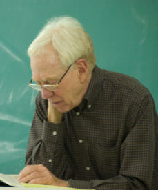 A photo of Dreyfus looking down at a paper with a green chalk board in the background.