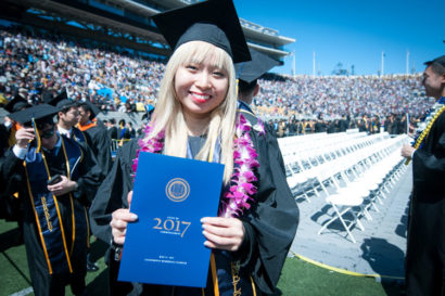 A photo of a UC Berkeley student standing outside in their graduation attire in the middle of a stadium in front of many rows of white chairs. 