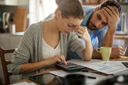 A stock photo of a stressed women sitting at a table using a calculator and touching her upper nose with her hand, while a man stands next to her with his elbows on the table and hand on his forehead also showcasing signs of stress. 