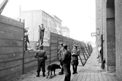 A black and white photo of guards along the East Berlin side of the Berlin Wall. They are two men on top of the gap in the wall with five others on the ground looking up at them.