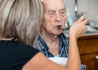 A generic stock photo of a lady spoon feeding an elderly man inside a room.