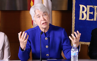 A photo of Carol Christ in the middle of speaking, with both her hands raised. She is sitting down at the conference panel table and the Berkeley logo can be seen on her right. 