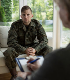 A photo of a man wearing green military camoflauge material, sitting on a couch with his hands together as a man takes notes on a clipboard sitting opposite from him. 