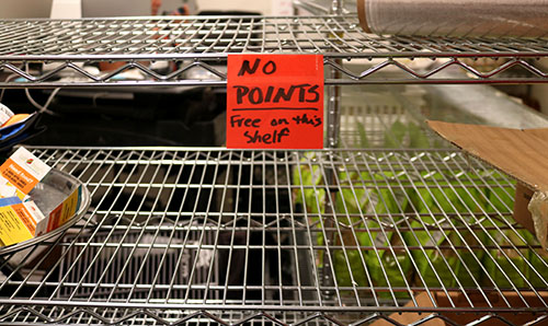 An empty metal shelf at a campus pantry with a sign that reads "NO POINTS, Free on this shelf," indicating free items that do not count toward the student limit.