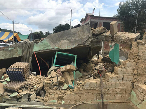 A collapsed adobe house in Totolapan, Morelos, following a recent earthquake, with debris and damaged furniture visible among the rubble.