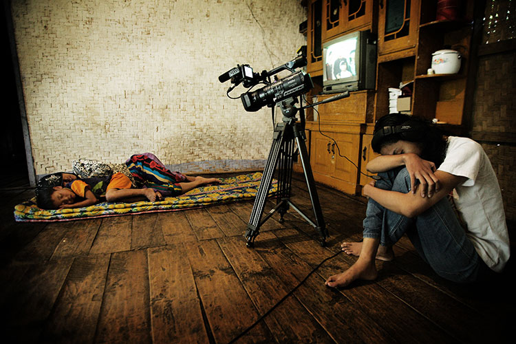 A video camera captures two children sleeping on the floor while a woman rests nearby, part of the Global Lives Project documenting daily life worldwide.