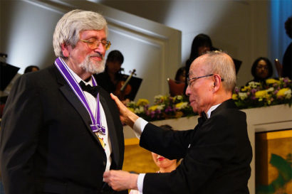 Taruskin wears a tuxedo and smiles as Imura places a medal around his neck during a ceremony. 