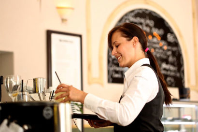 A women wearing a white button up shirt and black vest grabs a drink to serve to table at a restaurant.