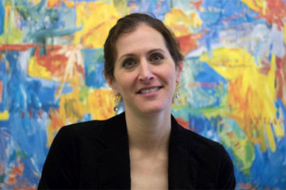 Amy Lerman wears a black blazer and sits in front of a bright, abstract painting.  