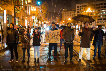A group of people stand in a side-by-side line on a street illuminated by city lights at night. One holds a sign that says "Black Lives Matter."