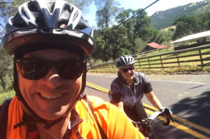 Selfie taken by a man wearing a helmet and sunglasses and on a bike with a woman on a bike coming up behind him.