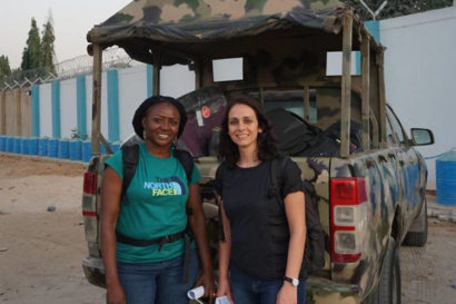 Grace Oyenubi and Nani Walker stand in front of pickup truck outfitted with a camouflage paint job and canopy top. Behind them is a tall wall adorned with razor wire
