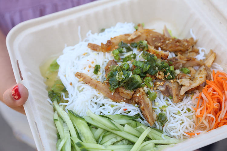 Seasoned pork on a bed of white noodles in a to-go box, garnished with cucumber slices, green onions and thinly peeled carrots