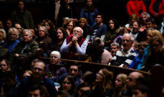 A crowd of mourners sit in a pew and clasp their hands in prayer. 