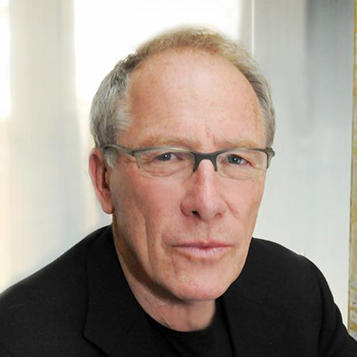 headshot of a person with short whitish hair wearing glasses and a black jacket with a neutral look on their face