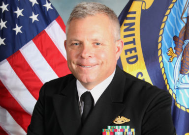 Capt. Scott Shackleton wears a suit displaying his military medals, with the American and US Navy flags in the background. 