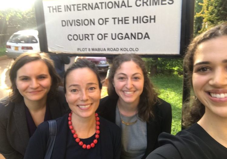A photo of four women smiling behind a sign that reads "The International Crimes Division of the High Court of Uganda" outside on a sunny day.