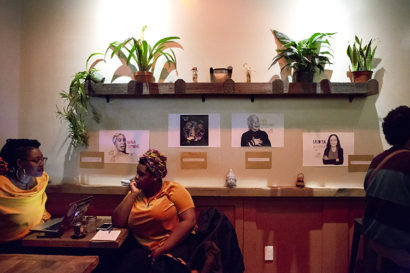 Another image inside the aforementioned cafe of the wall decorated with plants, wooden shelving, and photos of people as two people sit under it fully conversing. 