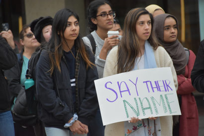 An image of a crowd of ten people standing solemnly while one person holds a sign that reads "say their name" in a bold fashioni.