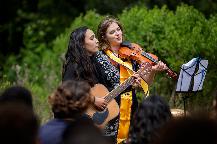 Two women stand together, one playing guitar and the other playing violin.