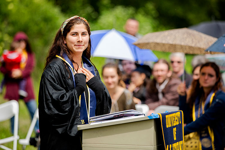 Elise Dimick stands at a podium outside with her hands on her heart during alternative graduation