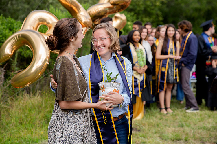 Skye Michel hands a small potted plant to a student in front of balloon banners during the alternative graduation ceremony