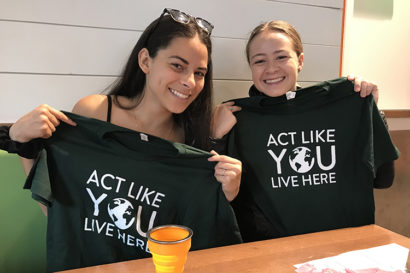 Sage and another female student hold up t-shirts that say "Act like you live here" with a picture of the earth