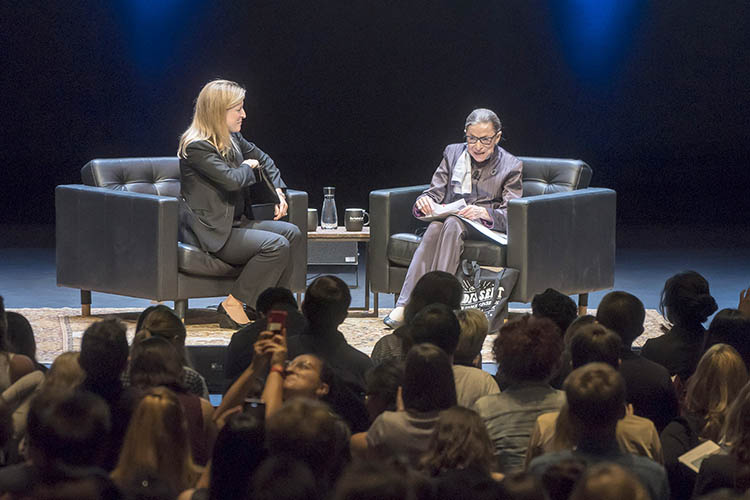 ginsburg talks on stage while sitting in a black chair