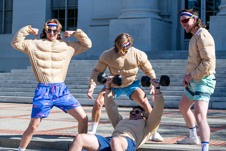 four students in puffy bobybuilding costumes pretend to lift heavy weights