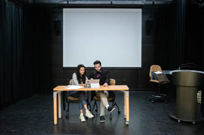 Students Salwa Meghjee and Drew Woodson sit at a table on stage, going over a script