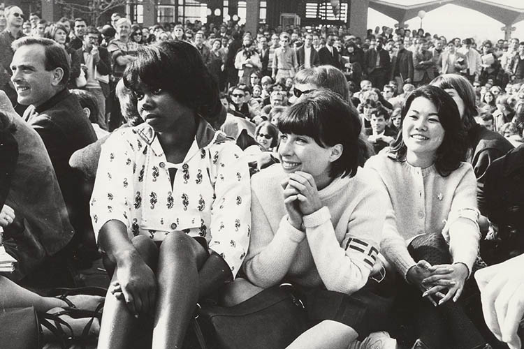 Three women sit on the ground listening to a speaker amidst a crowd of people.