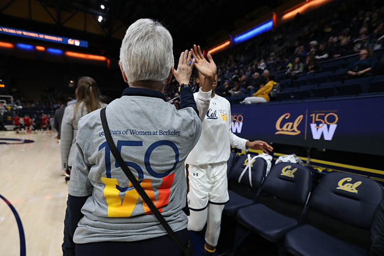 The back of a women wearing a grey t-shirt with the 150W logo faces the camera as she gives a high five to a basketball player.