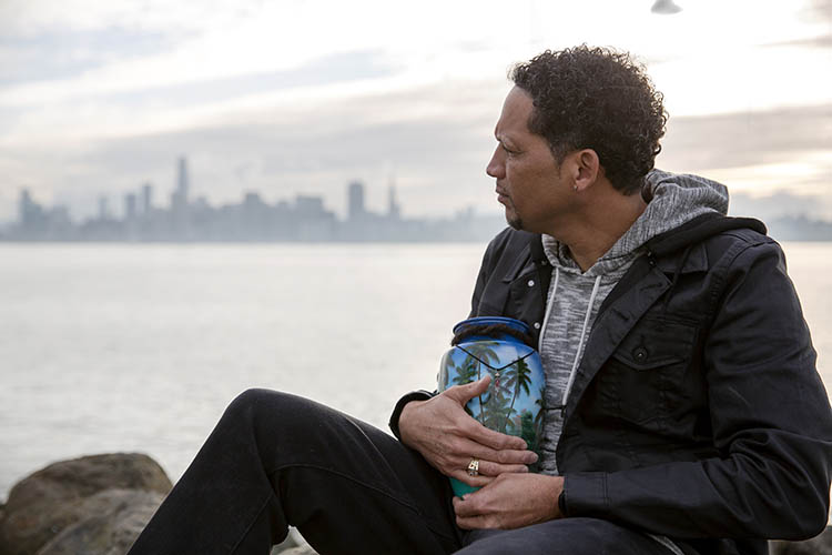 Richard Livingston Jr., whose son Richard Dejion Livingston III was murdered in 2015, sits outside cradling the urn that contains his son's remains. The sky is wintry and cool, and the skyline of San Francisco is in the background. (UC Berkeley photo by Brittany Hosea-Small)