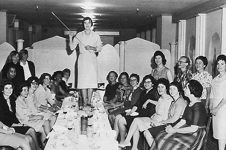 A women stands on a table holding a stick. She is surrounding by women sitting at her sides.