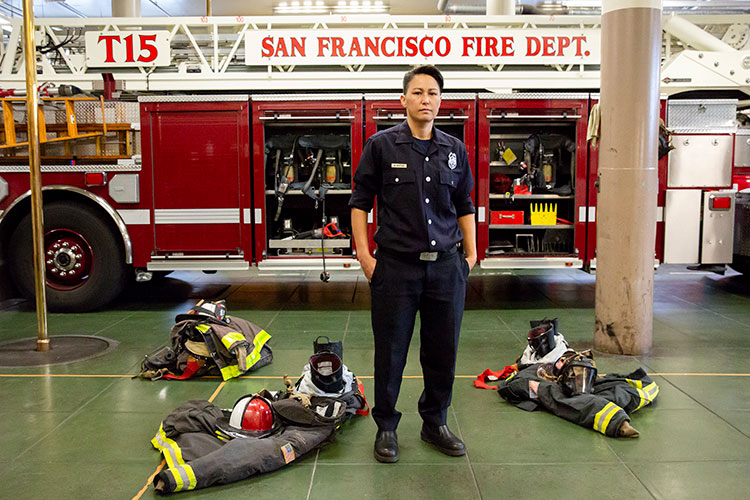 A photo of Maiko Bristow, a women firefighter with the San Francisco Fire Department. Maiko is wearing a black uniform and her turn out gear sits on the ground around her. Behind her is a fire truck with the words 