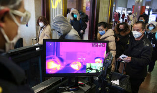 In the foreground, a person wearing a surgical face mask looks at a purple computer screen. In the background, people in a busy station, many wearing mask, line up.