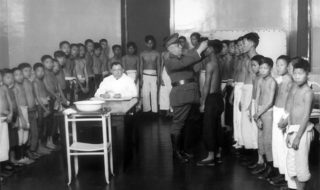 A group of men and boys without shirts on in a line as a man with a military uniform inspects them.