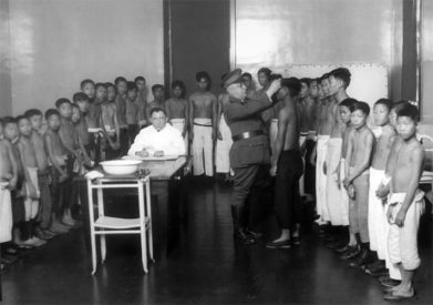 A group of men and boys without shirts on in a line as a man with a military uniform inspects them.