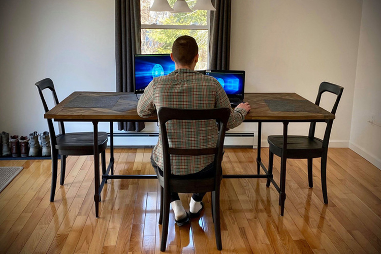 A worker practicing social distancing during the COVID-19 crisis works alone in a home dining room before two computer screens.