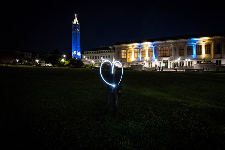 Sather Tower and Doe Library lit up in blue and gold while someone traces a heart with a flashlight in the foreground