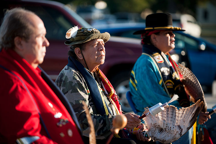 A photo of three men sitting outside at dusk. Two are wearing traditional Native American regalia.