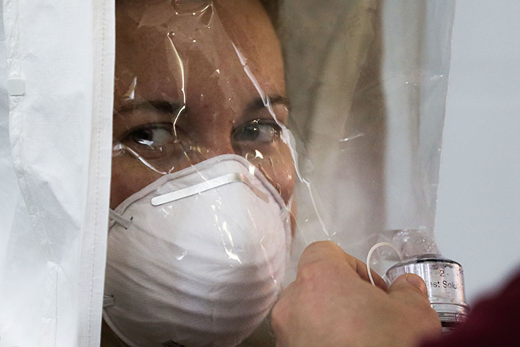 A photo of a person's face behind a white and clear plastic cover. The person is wearing a white face mask.