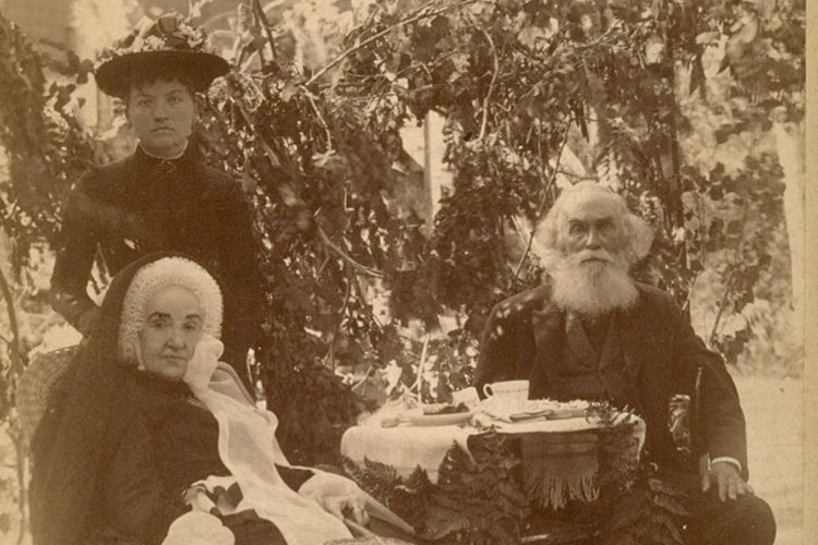Former UC President John LeConte, shortly before his death in 1891, is seated next to his wife, Eleanor and a caregiver. John LeConte, a physicist, is sporting a thick white beard, balding head and formal dark three-piece suit. His wife is wearing a bonnet, and the caregiver is standing behind the wife in dark dress and hat.