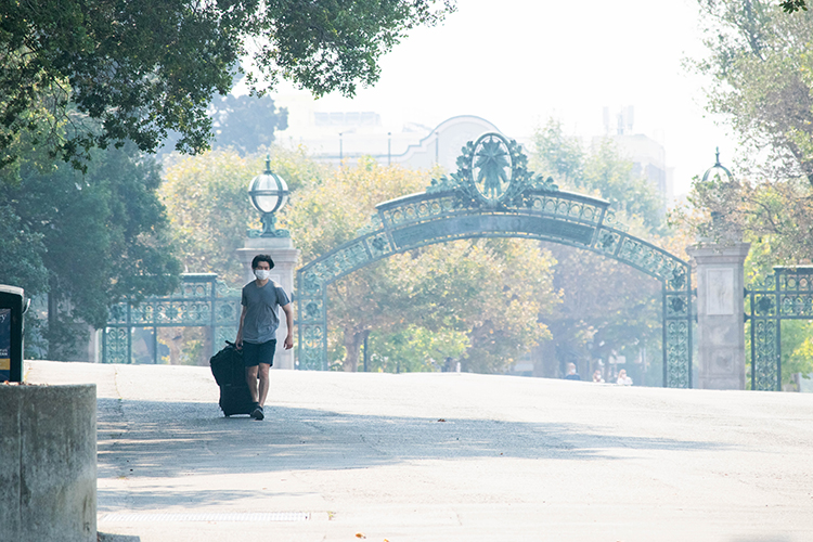 A student in a mask wheels his belongings onto campus near Sather Gate, with smoke from the Northern California wildfires visible in the air.