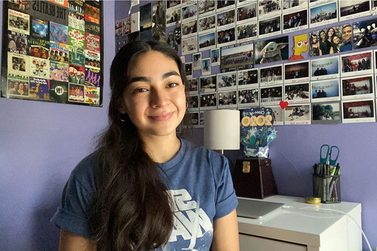 Incoming first-year student Daniela Castillo sits at her desk in her bedroom in Cathedral City, in California's Coachella Valley. The walls are purple and full of photos. She is smiling, sitting in a chair.